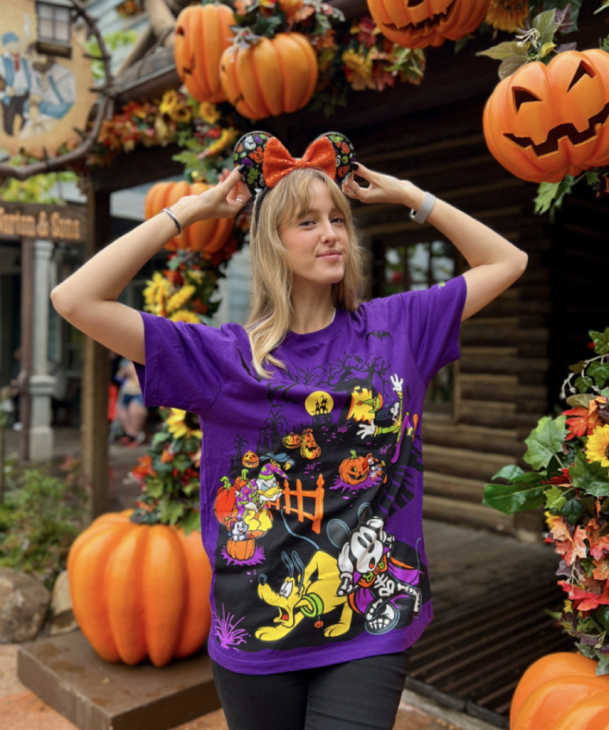 Une femme pose devant des décorations d'Halloween, vêtue d'une chemise violette avec des personnages de dessins animés et des oreilles de souris sur le thème d'Halloween, entourée de citrouilles et de feuilles d'automne. Sa brillante collection de badges d'échange de broches scintille sous la lumière du soleil d'automne, ajoutant un charme unique à la scène festive.