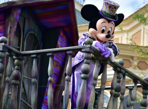 Un personnage en costume de Mickey Mouse s'appuie nonchalamment sur une balustrade en bois, arborant une tenue violette et verte vibrante avec un haut-de-forme. En arrière-plan, un bâtiment coloré se distingue par des détails complexes, rappelant le brouillon d'un artiste qui prend vie.