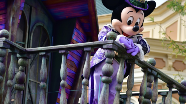 Un personnage en costume de Mickey Mouse s'appuie nonchalamment sur une balustrade en bois, arborant une tenue violette et verte vibrante avec un haut-de-forme. En arrière-plan, un bâtiment coloré se distingue par des détails complexes, rappelant le brouillon d'un artiste qui prend vie.