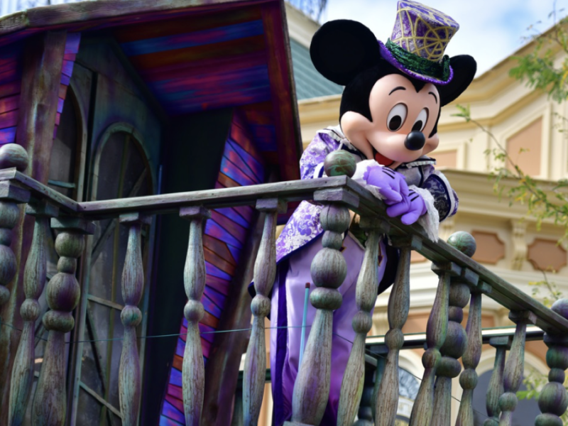 Un personnage en costume de Mickey Mouse s'appuie nonchalamment sur une balustrade en bois, arborant une tenue violette et verte vibrante avec un haut-de-forme. En arrière-plan, un bâtiment coloré se distingue par des détails complexes, rappelant le brouillon d'un artiste qui prend vie.