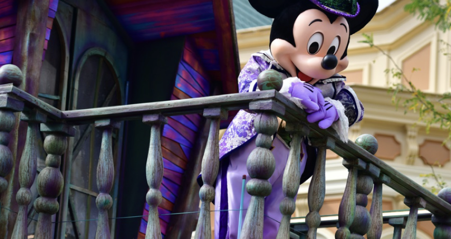 Un personnage en costume de Mickey Mouse s'appuie nonchalamment sur une balustrade en bois, arborant une tenue violette et verte vibrante avec un haut-de-forme. En arrière-plan, un bâtiment coloré se distingue par des détails complexes, rappelant le brouillon d'un artiste qui prend vie.