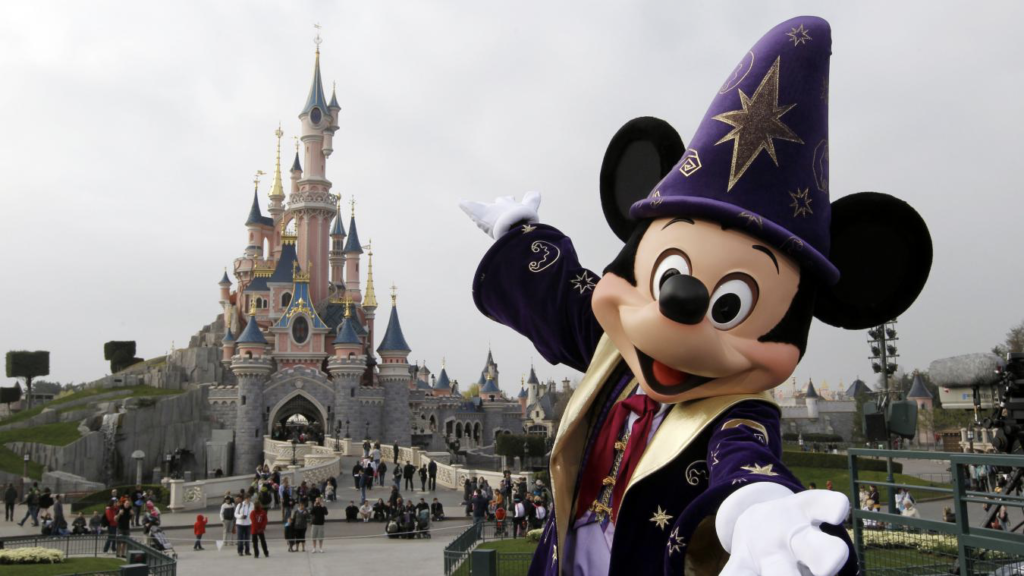 Un personnage en costume de sorcier Mickey Mouse pose joyeusement devant le château enchanteur de Disney, tandis que les visiteurs se promènent. Le ciel couvert crée un arrière-plan paisible, évoquant une sensation magique de « Zip-a-Dee-Doo-Dah ».