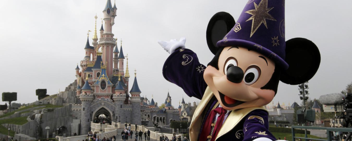 Un personnage en costume de sorcier Mickey Mouse pose joyeusement devant le château enchanteur de Disney, tandis que les visiteurs se promènent. Le ciel couvert crée un arrière-plan paisible, évoquant une sensation magique de « Zip-a-Dee-Doo-Dah ».