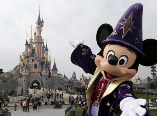 Un personnage en costume de sorcier Mickey Mouse pose joyeusement devant le château enchanteur de Disney, tandis que les visiteurs se promènent. Le ciel couvert crée un arrière-plan paisible, évoquant une sensation magique de « Zip-a-Dee-Doo-Dah ».