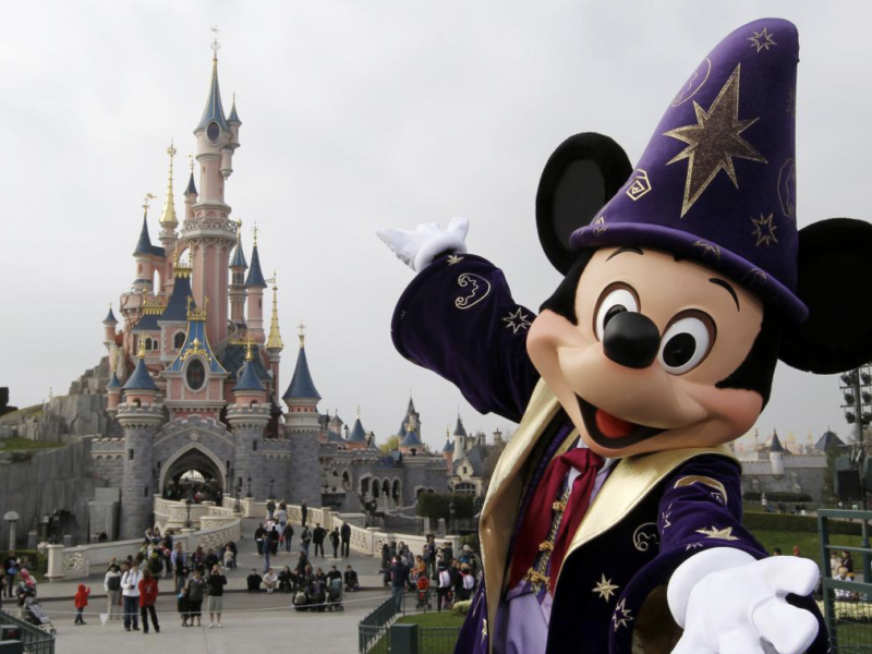 Un personnage en costume de sorcier Mickey Mouse pose joyeusement devant le château enchanteur de Disney, tandis que les visiteurs se promènent. Le ciel couvert crée un arrière-plan paisible, évoquant une sensation magique de « Zip-a-Dee-Doo-Dah ».