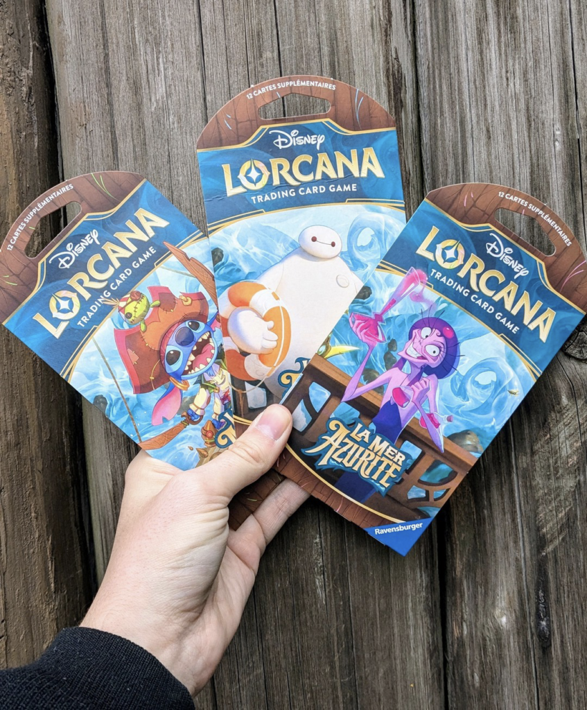 Une main tient trois packs de jeux de cartes à collectionner Disney Lorcana avec des illustrations colorées, rappelant des motifs d'échange d'épingles vibrants, sur un fond en bois texturé.