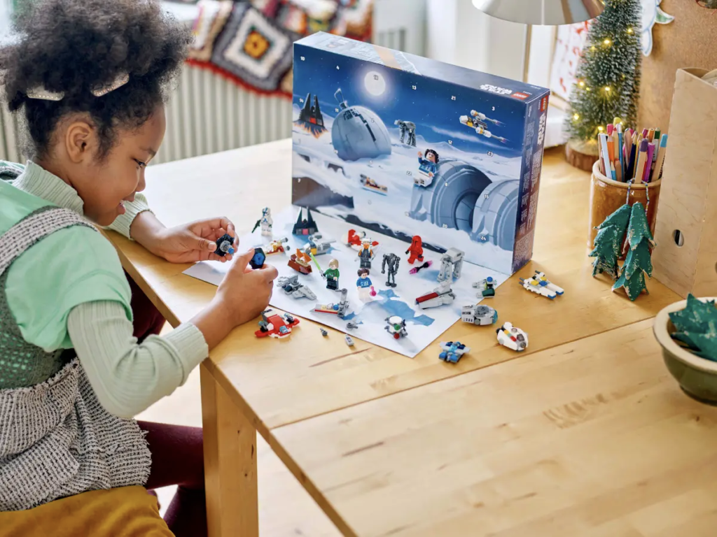 Un enfant joue avec enthousiasme avec un ensemble LEGO sur le thème de l'espace sur une table en bois, comprenant des mini figurines et des vaisseaux spatiaux, à côté d'une boîte ouverte avec une illustration de paysage enneigé.