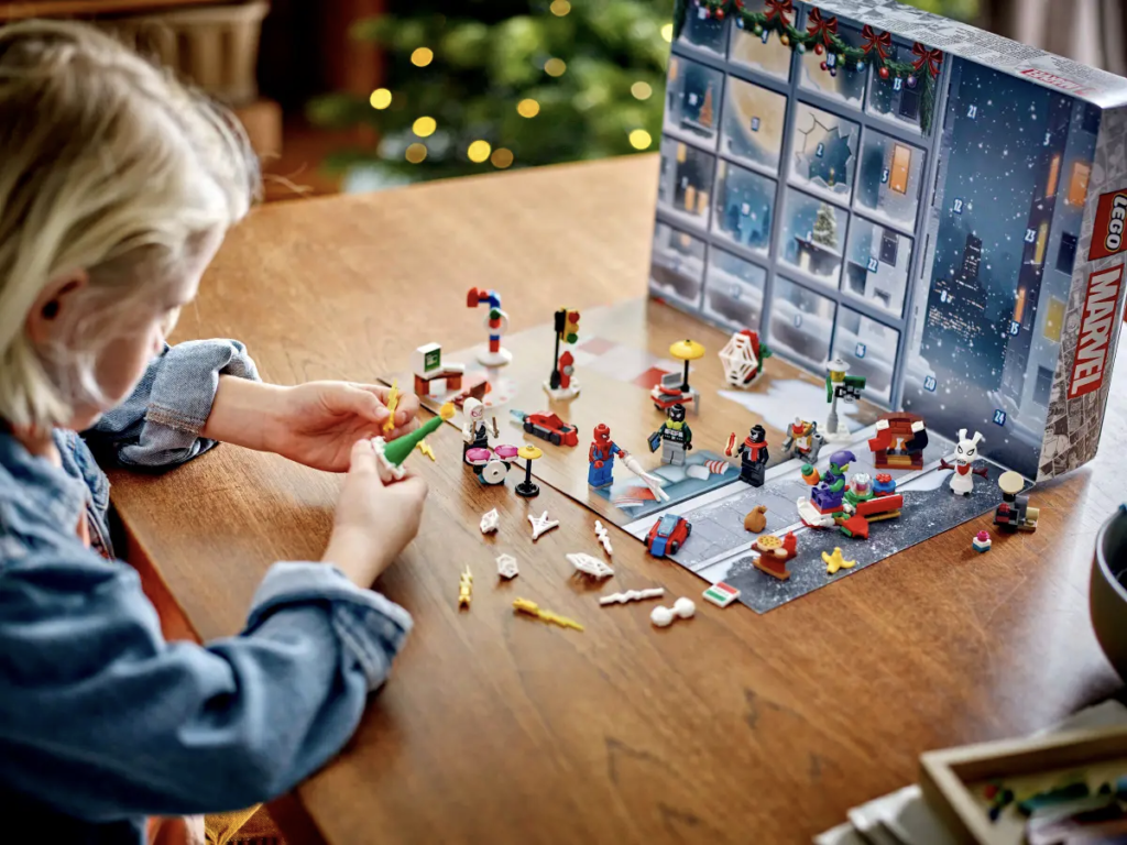 Un enfant assemble avec enthousiasme des pièces LEGO d'un calendrier de l'Avent sur le thème de Marvel sur une table en bois, entouré de la lueur festive du sapin de Noël, tout en observant sa collection d'échange de broches à proximité.