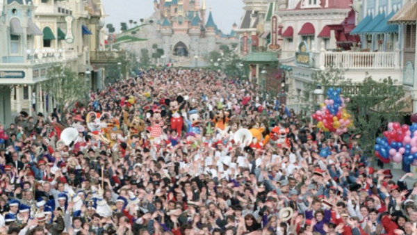 Une foule nombreuse s'est rassemblée dans la rue d'un parc à thème, avec costumes et décorations menant vers un château lointain, rappelant l'ouverture magique de Disneyland Paris le 12 avril 1992.