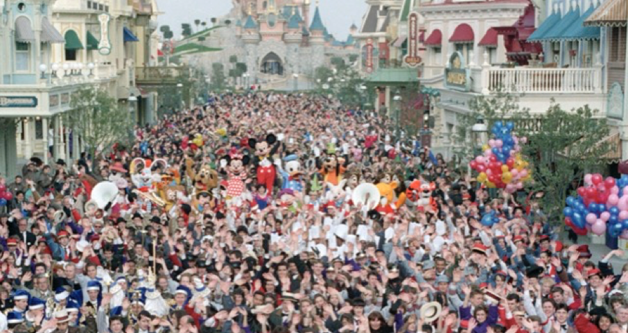 Une foule nombreuse s'est rassemblée dans la rue d'un parc à thème, avec costumes et décorations menant vers un château lointain, rappelant l'ouverture magique de Disneyland Paris le 12 avril 1992.
