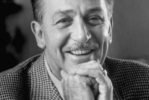 Un homme à moustache, vêtu d'un costume à carreaux, pose son menton sur ses mains jointes et sourit. Son attitude évoque le charme d'une époque qui rappelle la naissance de Walt Disney en 1901.