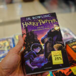 Dans une charmante librairie, une personne serre dans ses bras le livre « Harry Potter à l'école des sorciers », orné d'une étiquette de réduction de 25 %. Non loin de là, un fascinant brouillon de titres attend d'être découvert, chacun promettant de nouvelles aventures aux lecteurs avides.