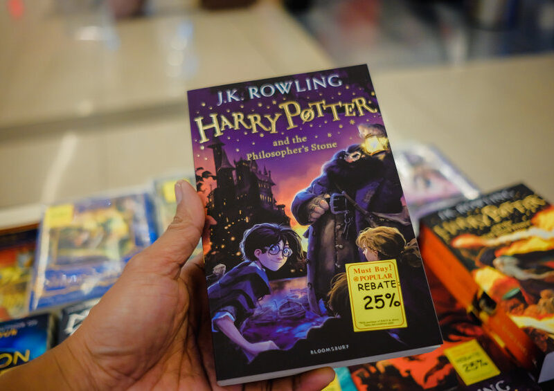 Dans une charmante librairie, une personne serre dans ses bras le livre « Harry Potter à l'école des sorciers », orné d'une étiquette de réduction de 25 %. Non loin de là, un fascinant brouillon de titres attend d'être découvert, chacun promettant de nouvelles aventures aux lecteurs avides.