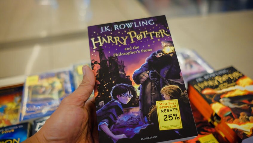 Dans une charmante librairie, une personne serre dans ses bras le livre « Harry Potter à l'école des sorciers », orné d'une étiquette de réduction de 25 %. Non loin de là, un fascinant brouillon de titres attend d'être découvert, chacun promettant de nouvelles aventures aux lecteurs avides.