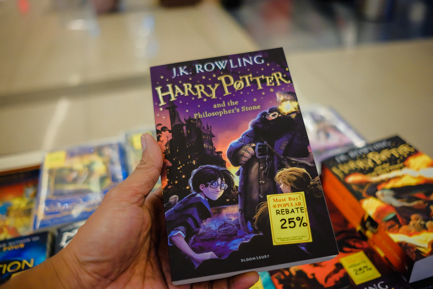 Dans une charmante librairie, une personne serre dans ses bras le livre « Harry Potter à l'école des sorciers », orné d'une étiquette de réduction de 25 %. Non loin de là, un fascinant brouillon de titres attend d'être découvert, chacun promettant de nouvelles aventures aux lecteurs avides.