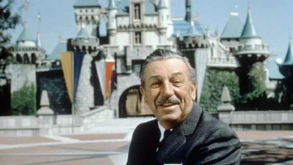 Un homme en costume se tient devant un château à Walt Disney World, souriant à la caméra.