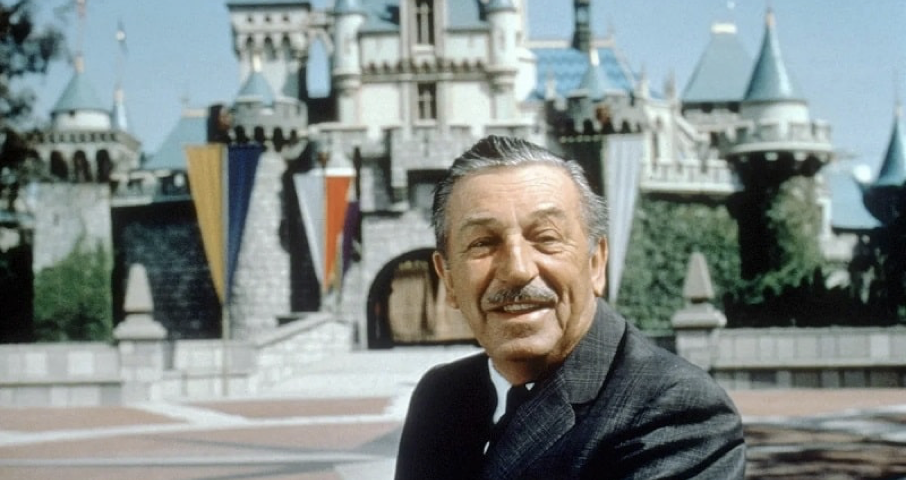Un homme en costume se tient devant un château à Walt Disney World, souriant à la caméra.