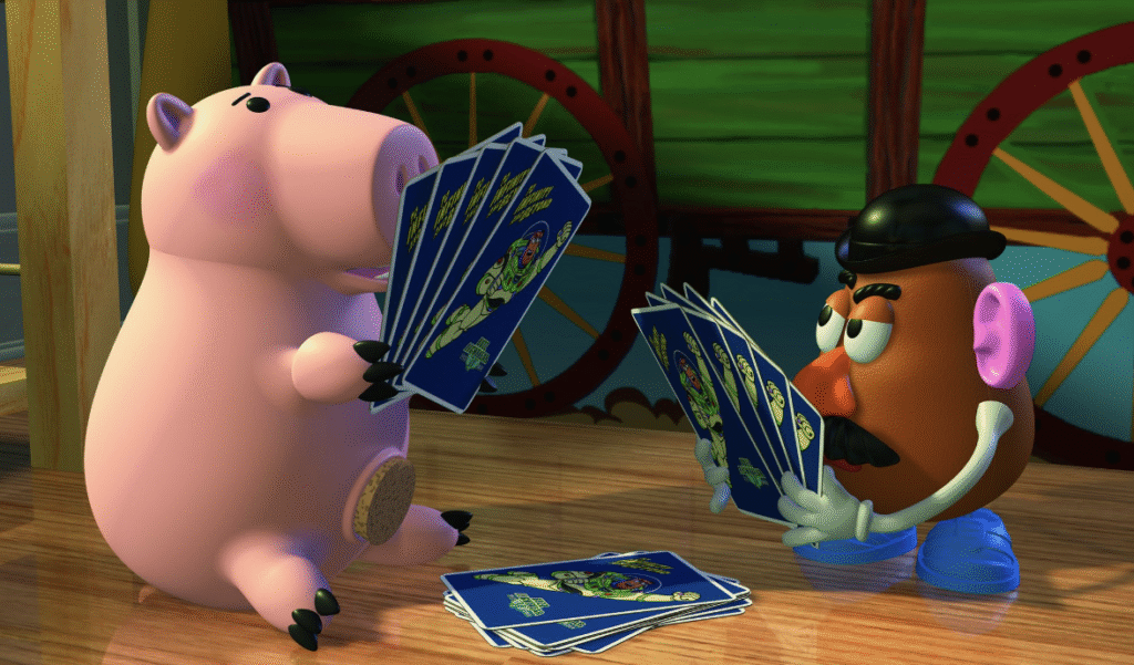 Une tirelire et une pomme de terre Toy Story sont posées sur un plancher en bois, tenant des cartes à jouer et se faisant face, avec des cartes éparpillées entre elles.