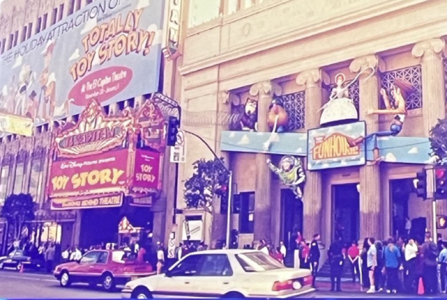 Vue de rue d'un théâtre décoré de bannières et de décorations vibrantes de Toy Story, d'une foule alignée à l'extérieur avec excitation et de plusieurs voitures passant sur la route.