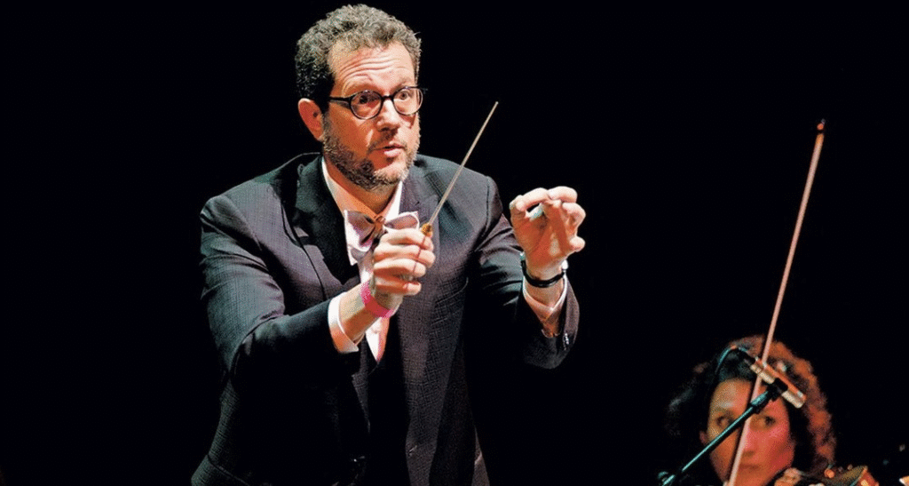 Un homme en costume et lunettes dirige un orchestre avec une baguette, tandis qu'un musicien joue d'un instrument à cordes en arrière-plan lors d'une représentation de "Le Festin".