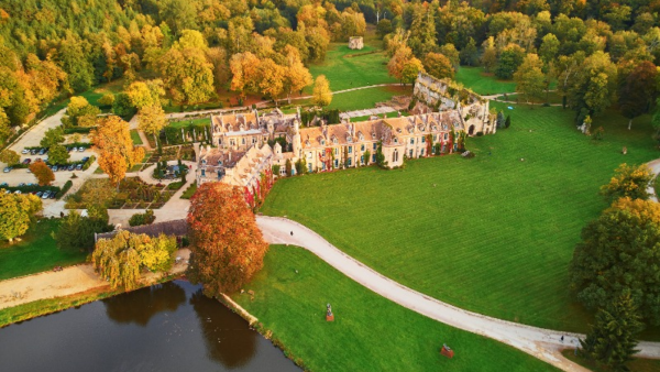 Vue aérienne d'un grand bâtiment historique entouré de pelouses vertes, d'arbres au feuillage d'automne, d'un étang et de sentiers sinueux dans un paysage rural, capturé comme une auto de Brouillon pour une inspiration scénique.