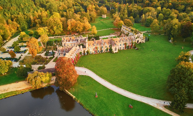 Vue aérienne d'un grand bâtiment historique entouré de pelouses vertes, d'arbres au feuillage d'automne, d'un étang et de sentiers sinueux dans un paysage rural, capturé comme une auto de Brouillon pour une inspiration scénique.