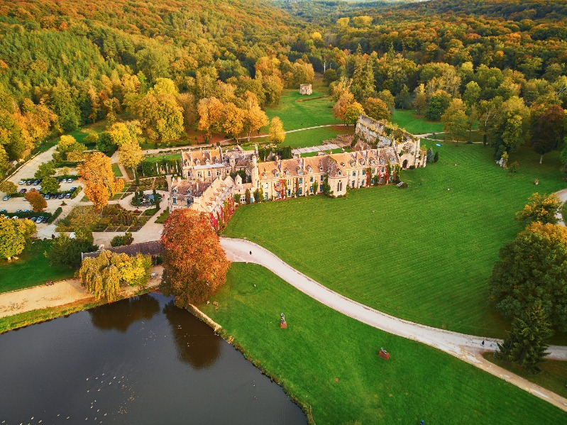 Vue aérienne d'un grand bâtiment historique entouré de pelouses vertes, d'arbres au feuillage d'automne, d'un étang et de sentiers sinueux dans un paysage rural, capturé comme une auto de Brouillon pour une inspiration scénique.
