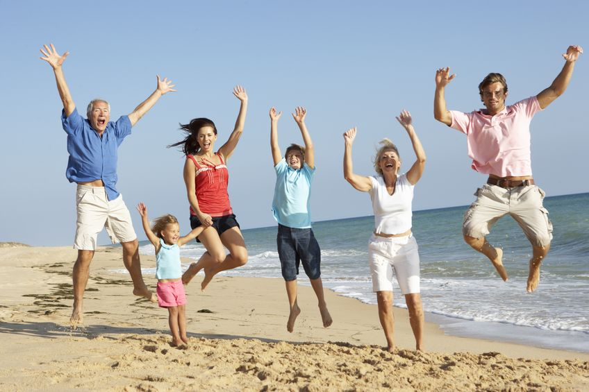 Six personnes et un enfant, issus de plusieurs générations, sautent dans les airs sur une plage de sable avec l'océan et le ciel bleu derrière eux, partageant des expériences uniques lors de leur séjour loin des Yvelines.