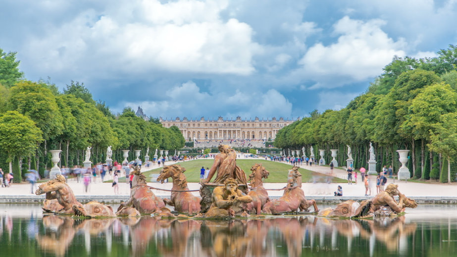 La fontaine d'Apollon dans les jardins du château de Versailles, Yvelines, avec le bâtiment du château en arrière-plan et les visiteurs profitant d'expériences uniques le long des allées.