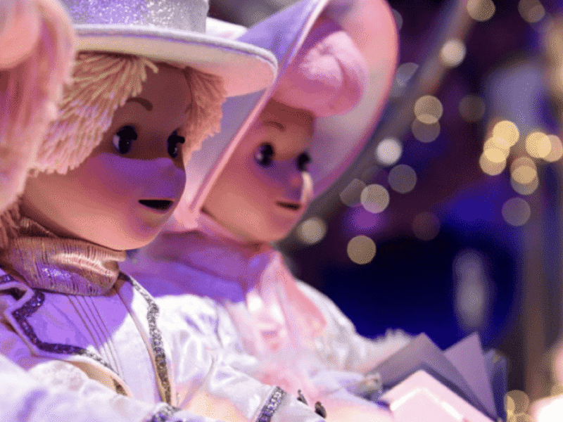 Gros plan de deux poupées vêtues de costumes et de chapeaux blancs, tenant des livres, avec de douces lumières violettes et jaunes en arrière-plan, évoquant une scène fantaisiste inspirée du style signature de Mary Blair.