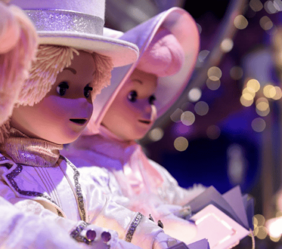 Gros plan de deux poupées vêtues de costumes et de chapeaux blancs, tenant des livres, avec de douces lumières violettes et jaunes en arrière-plan, évoquant une scène fantaisiste inspirée du style signature de Mary Blair.