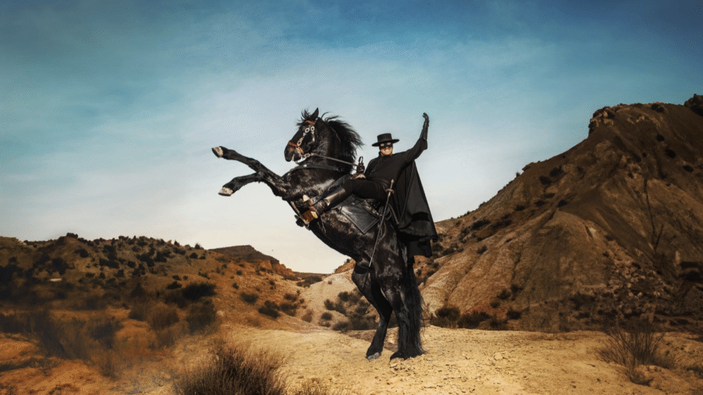 Un homme déguisé en Zorro, portant une cape et un chapeau noirs, monte un cheval noir cabré dans un paysage sec et vallonné sous un ciel clair.