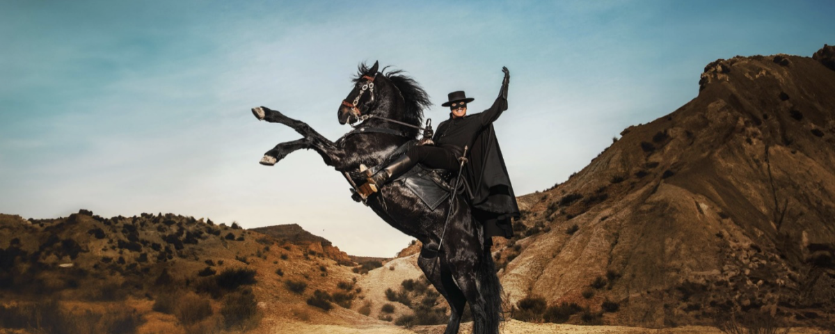 Un homme déguisé en Zorro, portant une cape et un chapeau noirs, monte un cheval noir cabré dans un paysage sec et vallonné sous un ciel clair.
