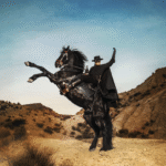 Un homme déguisé en Zorro, portant une cape et un chapeau noirs, monte un cheval noir cabré dans un paysage sec et vallonné sous un ciel clair.