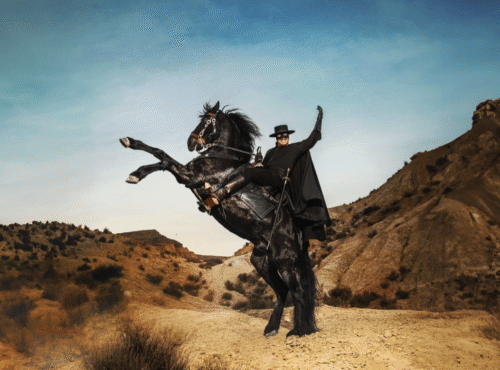 Un homme déguisé en Zorro, portant une cape et un chapeau noirs, monte un cheval noir cabré dans un paysage sec et vallonné sous un ciel clair.