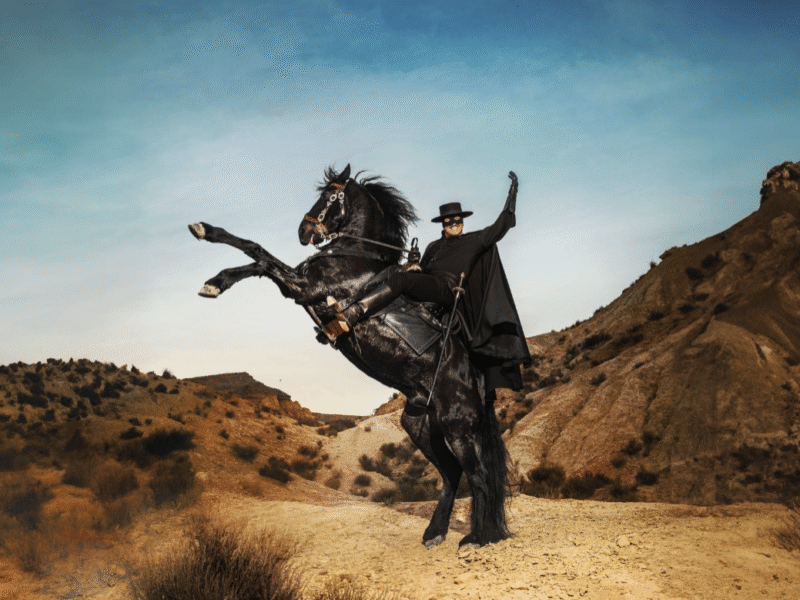 Un homme déguisé en Zorro, portant une cape et un chapeau noirs, monte un cheval noir cabré dans un paysage sec et vallonné sous un ciel clair.