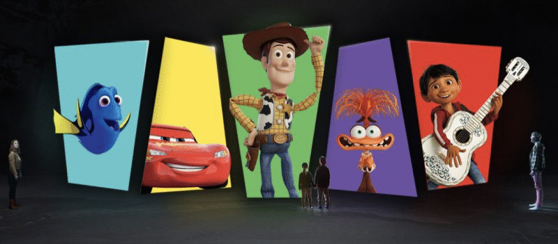 Cinq personnes se tiennent face à de grands écrans affichant les personnages bien-aimés de Pixar : Dory, Flash McQueen, Woody, une créature ressemblant à Muppet et un garçon avec une guitare.