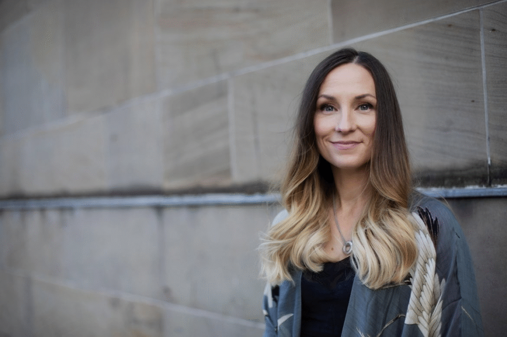 Une femme rebelle aux longs cheveux bruns et blonds se tient devant un mur de pierre beige, vêtue d'une veste à motifs gris et souriant légèrement à la caméra.