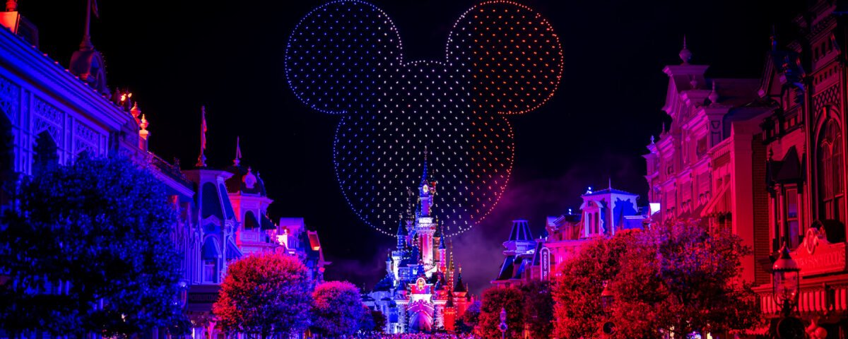 Une foule nombreuse se rassemble la nuit devant le château illuminé de Disneyland Paris, tandis que des drones et un feu d'artifice illuminent le ciel d'une tête de Mickey incandescente pour le 14 juillet 2025.
