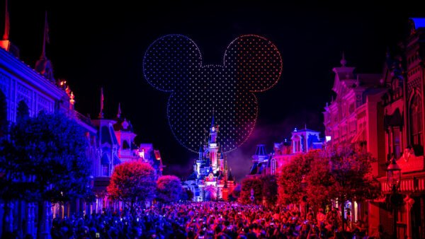 Une foule nombreuse se rassemble la nuit devant le château illuminé de Disneyland Paris, tandis que des drones et un feu d'artifice illuminent le ciel d'une tête de Mickey incandescente pour le 14 juillet 2025.