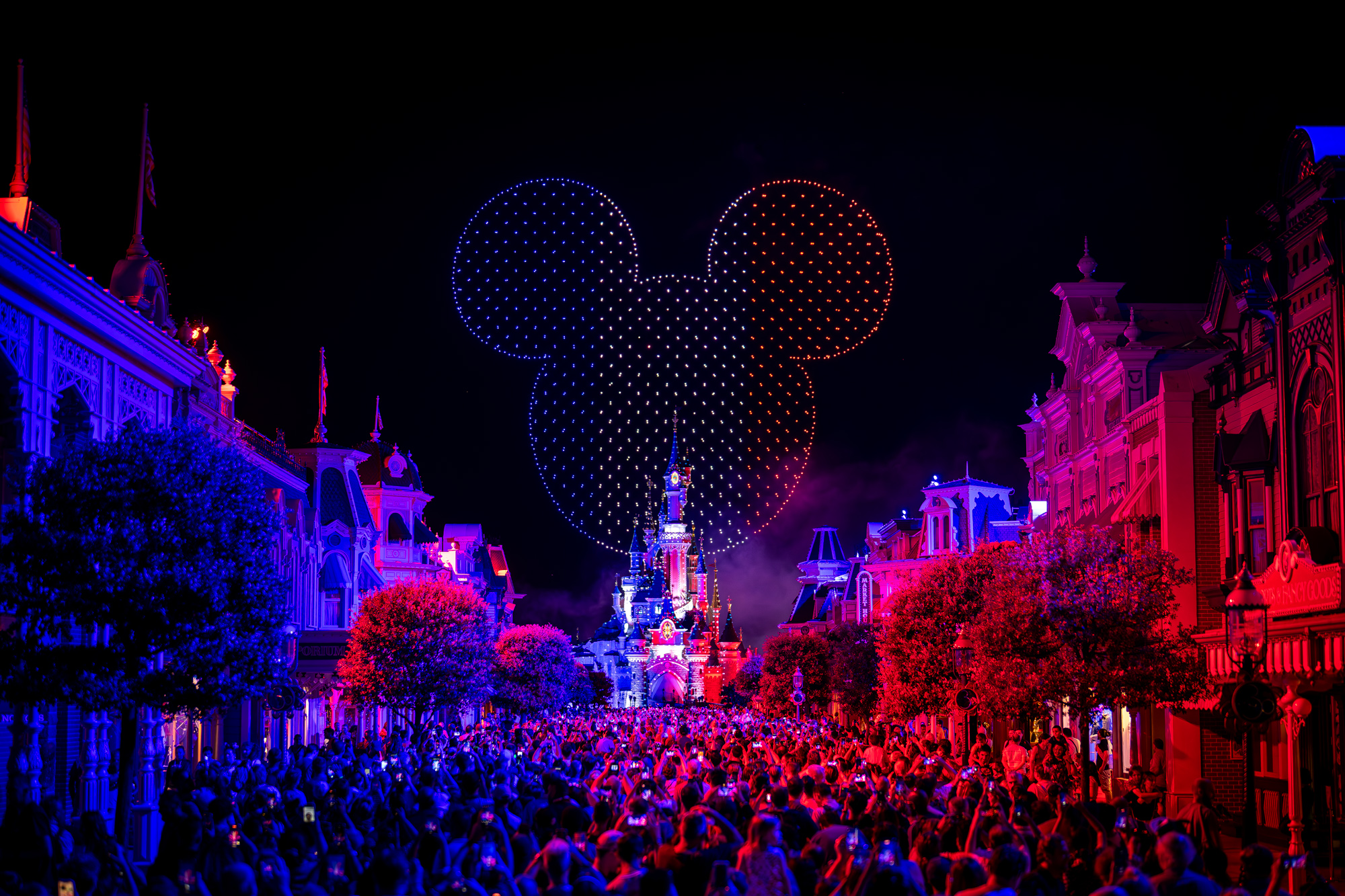 Une foule nombreuse se rassemble la nuit devant le château illuminé de Disneyland Paris, tandis que des drones et un feu d'artifice illuminent le ciel d'une tête de Mickey incandescente pour le 14 juillet 2025.