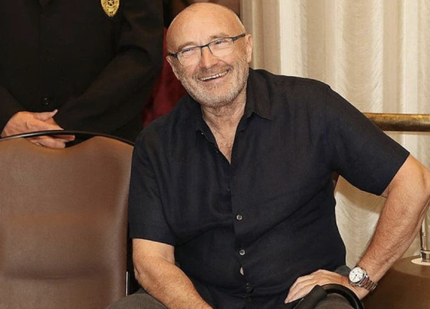 Un homme souriant, âgé, avec des lunettes et une chemise sombre, est assis à l'intérieur, posant une main sur une chaise et l'autre sur son genou, comme s'il se souvenait de Mon Frère Ours, un classique de la galaxie Disney.