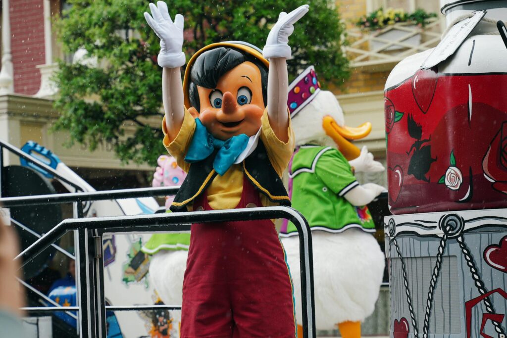 Une personne déguisée en Pinocchio lève les bras lors d'une parade de Disneyland Paris, avec un autre personnage déguisé en canard à l'arrière-plan.