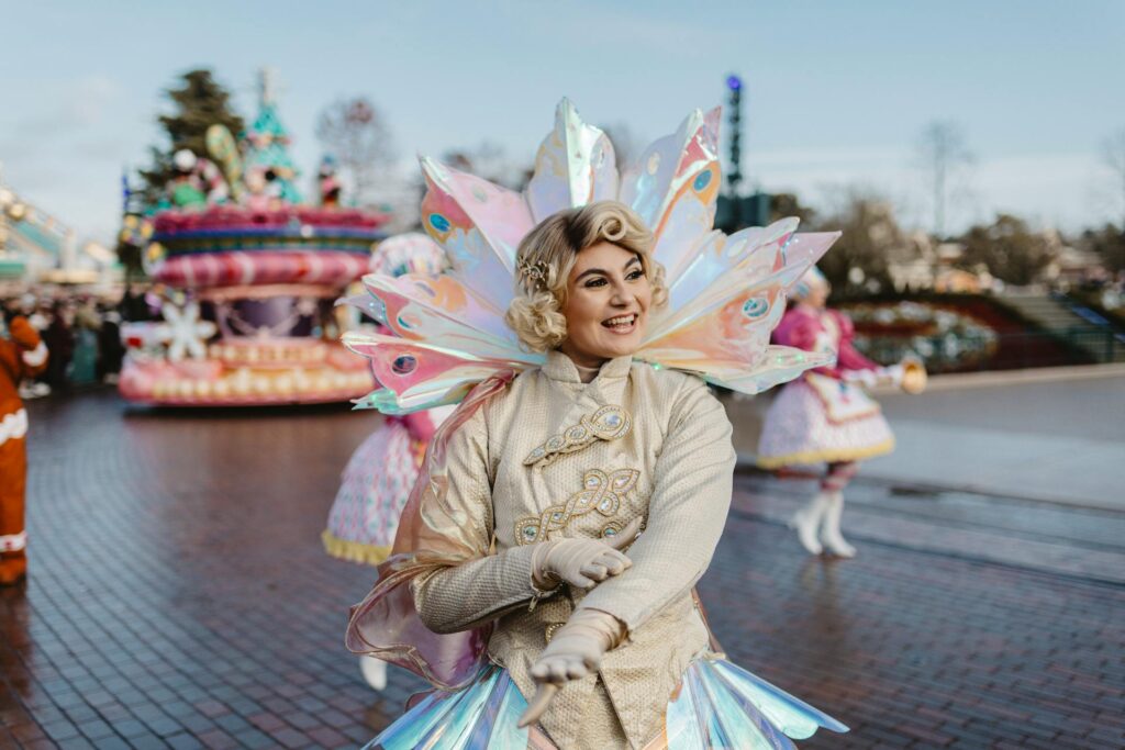 Un artiste en costume de fantaisie avec des ailes irisées sourit lors d'une parade à Disneyland Paris. Des chars et d'autres artistes sont visibles à l'arrière-plan tandis que les visiteurs profitent de la magie avec leurs billets.
