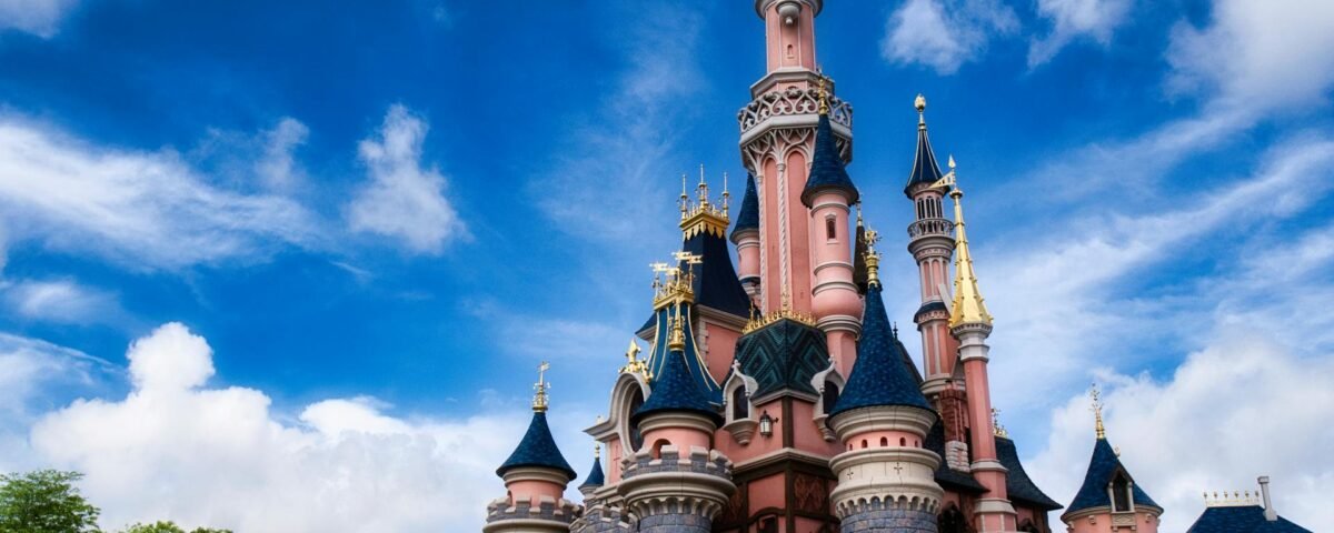 Un château de conte de fées rose et bleu avec des tours et des accents dorés se dresse sous un ciel bleu avec des nuages épars, invitant les visiteurs à vivre la magie de Disneyland Paris et à découvrir ses nouveaux billets à tarification dynamique.