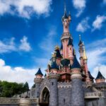 Un château de conte de fées rose et bleu avec des tours et des accents dorés se dresse sous un ciel bleu avec des nuages épars, invitant les visiteurs à vivre la magie de Disneyland Paris et à découvrir ses nouveaux billets à tarification dynamique.