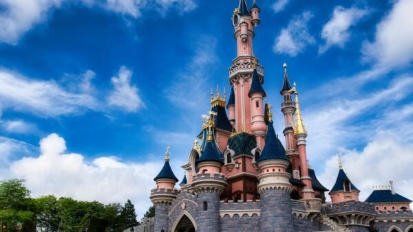 Un château de conte de fées rose et bleu avec des tours et des accents dorés se dresse sous un ciel bleu avec des nuages épars, invitant les visiteurs à vivre la magie de Disneyland Paris et à découvrir ses nouveaux billets à tarification dynamique.