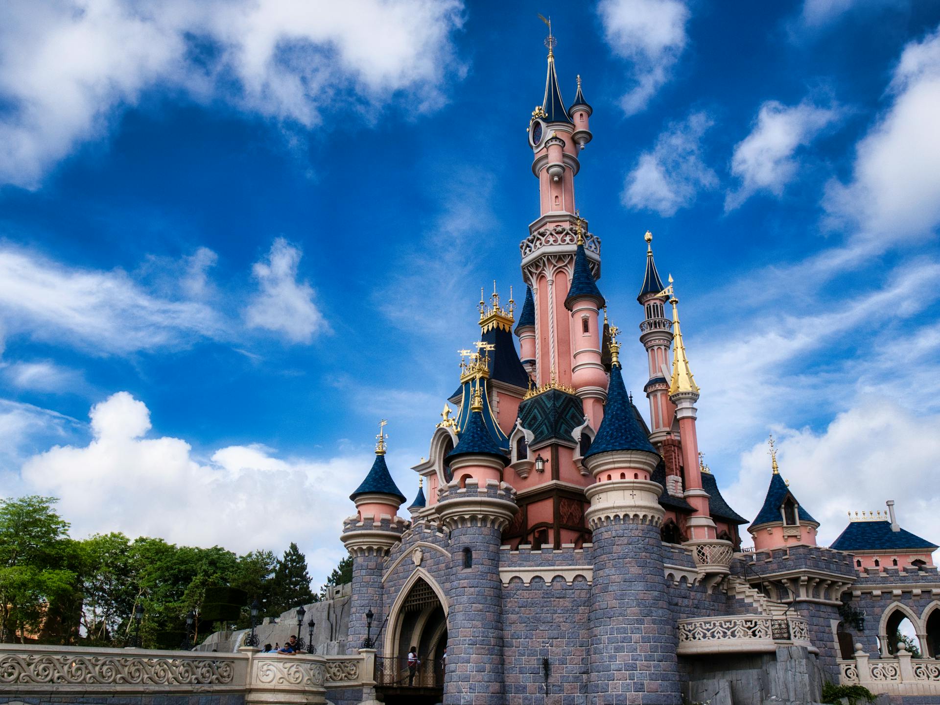 Un château de conte de fées rose et bleu avec des tours et des accents dorés se dresse sous un ciel bleu avec des nuages épars, invitant les visiteurs à vivre la magie de Disneyland Paris et à découvrir ses nouveaux billets à tarification dynamique.