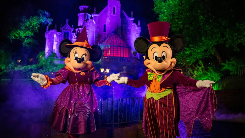 Mickey et Minnie Mouse portent des costumes d'Halloween et se tiennent devant un château illuminé la nuit pendant Halloween Disneyland, entourés de lumières colorées et de brouillard.