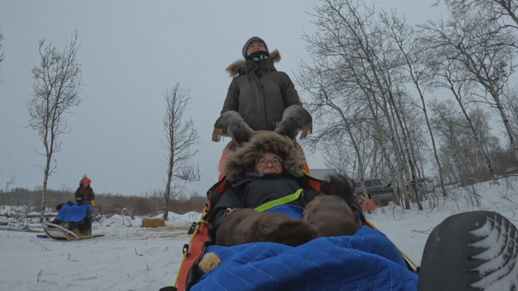 Deux personnes emmitouflées dans des vêtements d'hiver se déplacent en traîneau à chiens dans un paysage enneigé aux arbres dénudés et au ciel couvert, dans une scène digne d'une série documentaire du National Geographic.